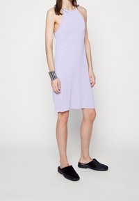 Vrouw in een mouwloze lavendelkleurige jurk tot de knie, zilveren armband en zwarte instapschoenen, staand tegen een witte achtergrond.