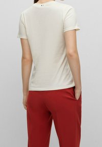 Camiseta corta de manga corta color crema con un escote redondo, textura suave y un pequeño detalle de logo en la parte de atrás, combinada con pantalones rojos.