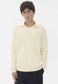 Világos sárga kötött pulóver V-nyakkal és hajtott gallérral, hosszú ujjakkal és bordázott mandzsettákkal, sötét nadrággal párosítva.