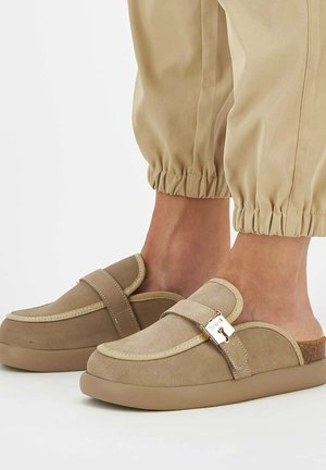 Beige ruskinds slip-on sko med en rund tå, en dekorativ rem og en kraftig gummisål. Tekstureret stofdetalje og metalornament.
