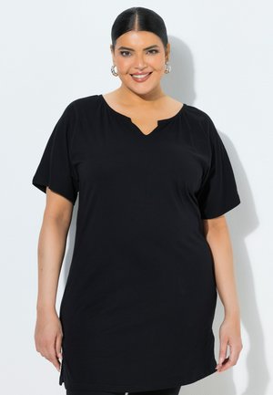 Uśmiechnięta kobieta o ciemnych włosach, w kolczykach-kołach, ubrana w czarną tunikę z krótkim rękawem i dekoltem w kształcie V na tle białego tła.