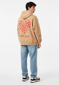 Bež hoodie s kapuljačom s velikim grafičkim motivom crvenog srca na leđima, natpisom "SWEET heart" i ležernim trapericama s izblijedjelim završetkom.