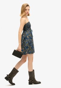 Robe sans bretelles bleu à motifs cachemire avec un corsage élastique et une taille à cordon de serrage, associée à des bottes en cuir marron jusqu'au genou et un sac à main noir en tissu tissé.