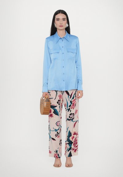 Cămașă butonată din satin de culoare albastră deschisă, cu două buzunare pe piept, asortată cu pantaloni largi cu imprimeu floral în nuanțe de crem, roz și teal. Geantă țesută din piele maro.