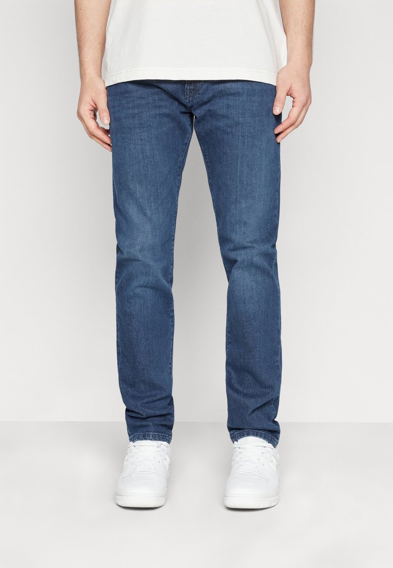 Homme portant un jean slim bleu et des baskets blanches, debout devant un fond clair uni, les mains détendues le long du corps.