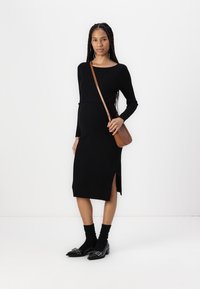 Černé žebrované šaty s dlouhými rukávy a bočním rozparkem, doplněné černými ponožkami a lesklými černými mokasíny. Hnědá crossbody taška.