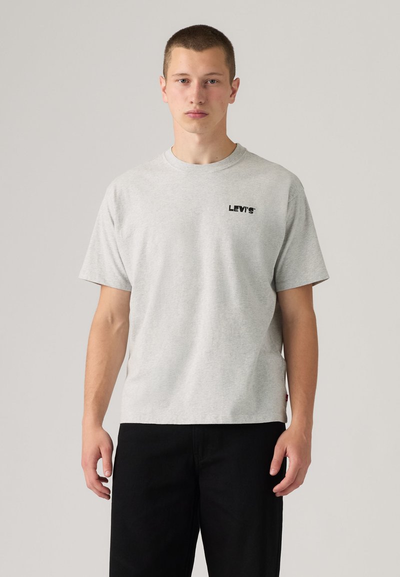 Junger Mann, der nach vorne blickt, trägt ein hellgraues Levi's T-Shirt und schwarze Hose vor einfachem Hintergrund.