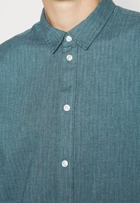 Camicia turchese con motivo a spina di pesce e colletto, decorata con bottoni bianchi sul davanti. Il tessuto ha una finitura morbida e testurizzata.