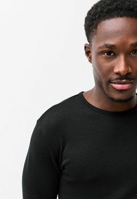 Čierne rebrované tričko s dlhým rukávom a okrúhlym výstrihom. Detailný záber ukazuje textúru a strih, doplnené neutrálnym pozadím.