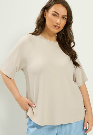 Kvinde med langt brunt hår iført lys beige løs kortærmet top og lyseblå jeans, poserer med hånd i lommen foran ensfarvet baggrund.