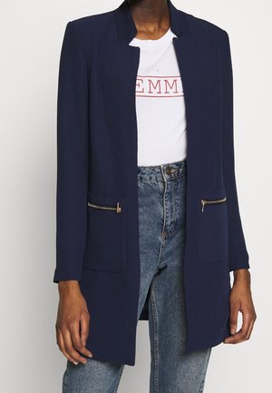Persoon draagt een donkerblauwe blazer met open voorkant en gouden ritssluitingen op de zakken over een wit t-shirt en blauwe spijkerbroek.