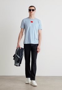 Tricou albastru deschis cu etichetă roșie, pantaloni negri și adidași albi. Ținând o jachetă din denim, purtând ochelari de soare negri. Design simplu, look casual.