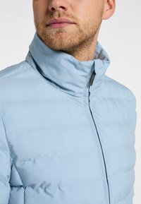 Giacca imbottita azzurro chiaro con colletto alto, texture liscia e chiusura con zip. Presenta cuciture orizzontali per l'isolamento.