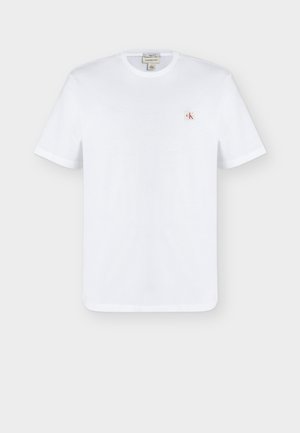 Biała koszulka z krótkim rękawem i okrągłym dekoltem z małą czerwoną i czarną naszywką z logo "ck" na lewej piersi.