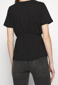 Mujer vistiendo una blusa negra de manga corta con detalle de cintura fruncida, combinada con jeans gris oscuro, vista desde la espalda contra un fondo blanco.