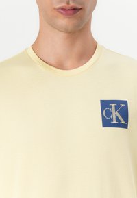 Camiseta de manga corta amarilla claro, hecha de una tela suave, con un logotipo cuadrado azul y letras blancas en la parte superior izquierda del pecho.