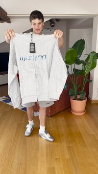 Világosszürke, cipzáros kapucnis pulóver, fényes kék "wasted" felirattal. Nagy ujjak jellemzik, és a címke még rajta van. A háttérben zöld növények találhatók.