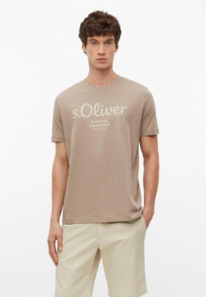 Jeune homme aux cheveux bruns courts, portant un t-shirt beige avec le logo "s.Oliver" et un short kaki clair, debout avec une main dans la poche.