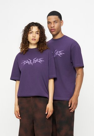 Twee modellen staan naast elkaar, gekleed in paarse grafische T-shirts en donkerbruine wijde broeken tegen een effen witte achtergrond.