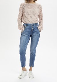 Lace long-sleeve top in soft blush, featuring textured floral patterns; paired with light blue high-waisted jeans with button accents. White sneakers.