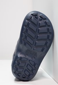 Suola di Crocs blu navy, caratterizzata da un motivo di battistrada testurizzato, forma curvata e logo in rilievo, con materiale in gomma liscia e dettagli delle cuciture.