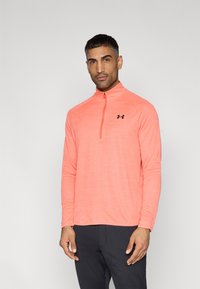 Μπλούζα με μακριά μανίκια και φερμουάρ σε ζωντανό κοραλί χρώμα, κατασκευασμένη από υφασμάτινη υφή με μαύρο λογότυπο στο στήθος. Απλός, αθλητικός σχεδιασμός.