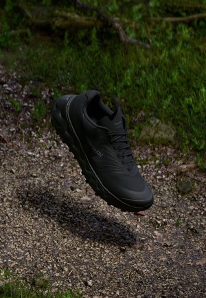 Adidas de alergare negre cu un superior texturat și o talpă amortizată, având lugs distinctive pentru tracțiune, prezentate pe un fundal natural.