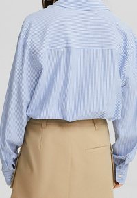 Bakåtsikt av en person som bär en blå och vit randig långärmad skjorta instoppad i hög midja beige byxor med bälteshällor.