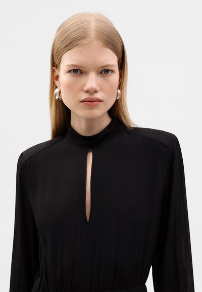 Czarna, długa sukienka z długim rękawem, wysokim kołnierzem i wycięciem w kształcie otworu przy szyi; gładka tekstura materiału; srebrne kolczyki w kształcie kół.