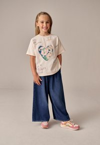 Tricou alb cu un design colorat în formă de inimă și text, asociat cu pantaloni închiși din denim albastru și adidași roz, crem și bej.