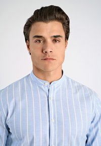 Camisa azul claro con rayas blancas verticales, cuello mandarín y cierre de botones. La tela parece suave con una textura delicada.