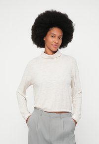 Woman with afro hairstyle wearing a light beige turtleneck sweater and gray pinstriped trousers, hands in pockets, smiling slightly.