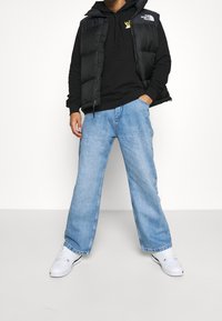 Gilet imbottito nero sopra una felpa nera con cappuccio, abbinati a jeans a gamba larga azzurri e sneakers bianche. Presenta loghi sul gilet e sulla felpa.