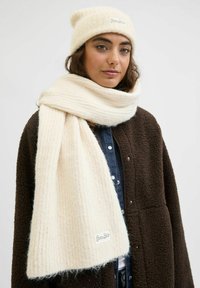 Kreemist kudumist beanie ja sobiv ribakaar, mis on valmistatud pehmest materjalist, koos pruunist fliisjakist ja teksapluusiga.