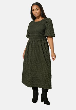 Vestido verde oscuro de longitud media con mangas abullonadas, corsé fruncido, patrón de cuadrícula texturizada, combinado con botas negras hasta la rodilla.