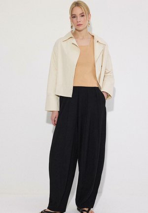 Vrouw met blond haar, goudkleurige oorringen, beige cropped jack, beige top, wijde zwarte broek en zwarte sandalen, staand tegen een witte muur.