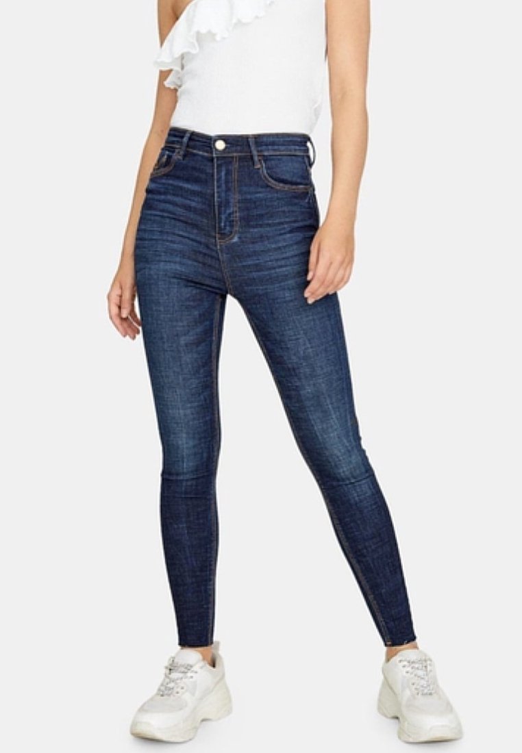 Hoge taille donkere blauwe skinny jeans met een gladde textuur en minimale vervaging, gecombineerd met witte sneakers en een gerimpeld wit topje.