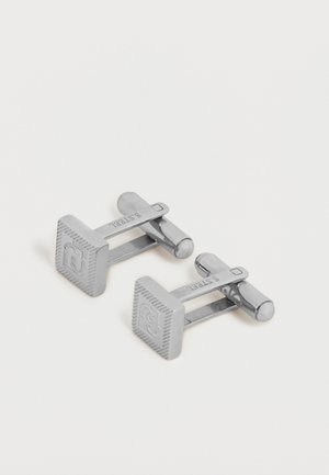 Een paar zilveren vierkante manchetknopen met gegraveerd logo en "S STEEL" gemarkeerd op de staaf, rustend op een witte ondergrond.