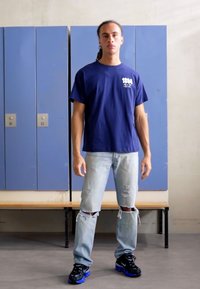 Navy cotton t-shirt with front graphic, distressed light blue denim jeans, and black sneakers with blue accents, standing against lockers.