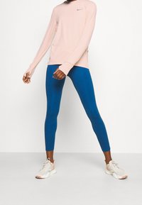 Helles rosa Langarmshirt mit Daumenlöchern, kombiniert mit blauen Leggings und weißen Sneakers mit korallenfarbigen Akzenten, vor einem neutralen Hintergrund.