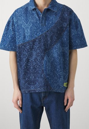 Mand iført kortærmet blå mønstret polo-shirt med buet mørkeblå design og blå jeans, stående foran en ensfarvet lys baggrund.