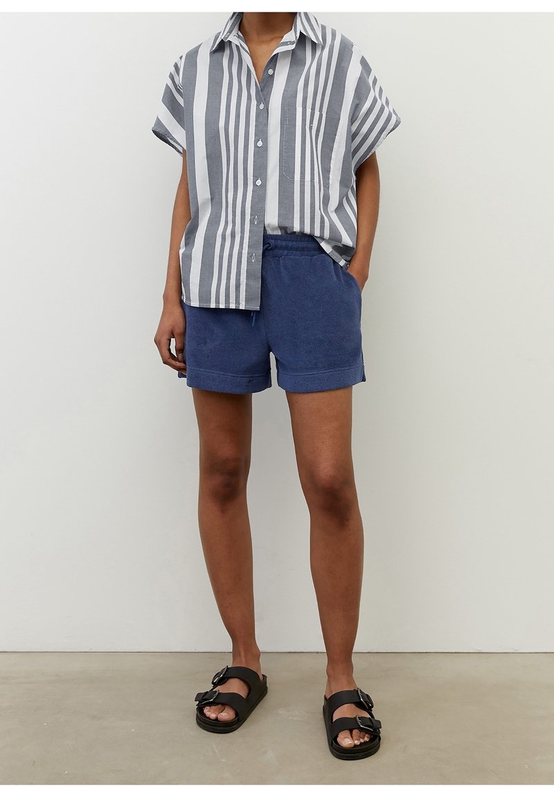 Personne portant une chemise à manches courtes rayée verticalement gris et blanc, un short bleu marine et des sandales noires à boucle, debout sur un sol gris.