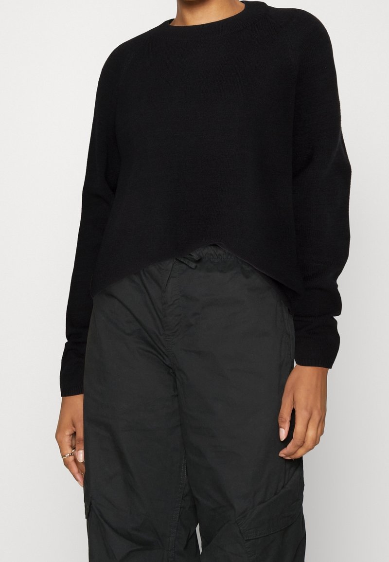 Femme portant un pull noir uni à manches longues et un pantalon cargo noir ample, debout les mains détendues le long du corps.