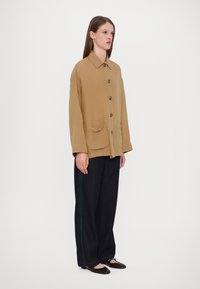 Tan, oversized button-up jacket with a pointed collar, front pockets, and brown buttons, paired with loose dark denim jeans and flats.