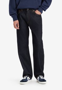 Personne portant un jean droit bleu foncé, des baskets bleu marine et blanches, et un sweat à capuche bleu marine, une main dans la poche.