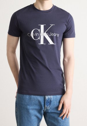 Homme portant un t-shirt Calvin Klein Jeans bleu foncé avec un grand logo blanc "CK" et un jean bleu clair sur un fond clair uni.