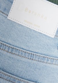 Tejido de mezclilla de color azul claro con una etiqueta blanca visible que presenta texto dorado. La etiqueta dice "Bershka ORIGINAL DENIM". El detalle de la costura es prominente.