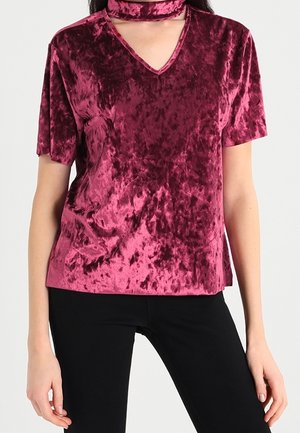 Frau trägt ein kurzärmeliges, burgunderfarbenes Samtoberteil mit V-Ausschnitt und angebrachter Halsbinde, kombiniert mit schwarzen Hosen.