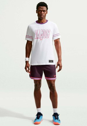 Man draagt een wit "VAMOS" t-shirt, donkere korte broek met rode strepen, sportpolsband, witte sokken en blauwe hardloopschoenen, staand op een effen achtergrond.