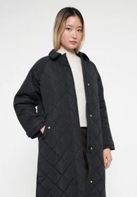 Quilted black long coat with a collar, snap buttons, and side pockets. The fabric has a smooth texture with diamond patterns.